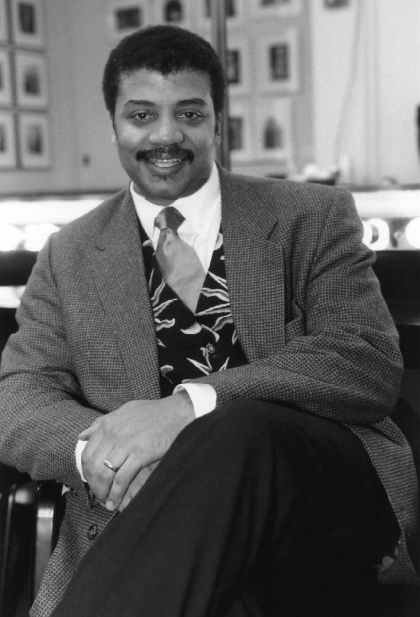 Neil deGrasse Tyson casually seated with crossed legs in a black and white portrait.