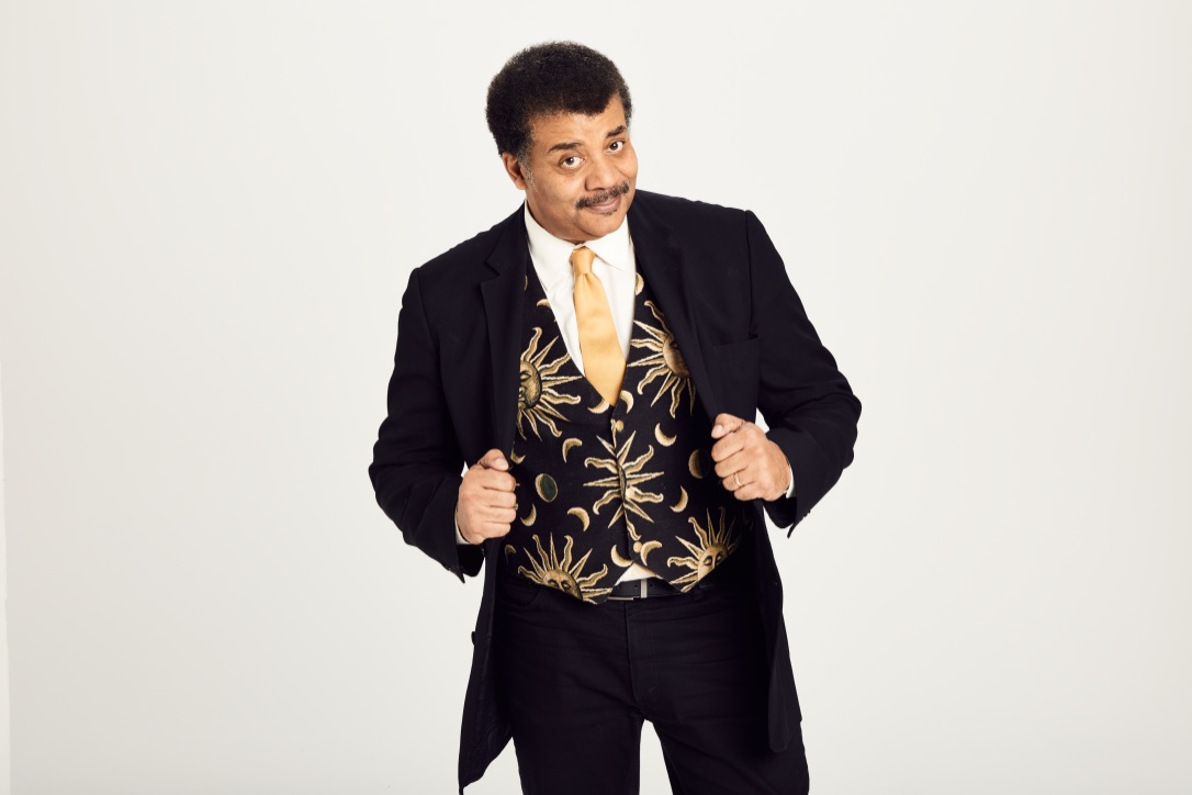 Neil deGrasse Tyson standing, leaning into the camera with an engaging look on his face.