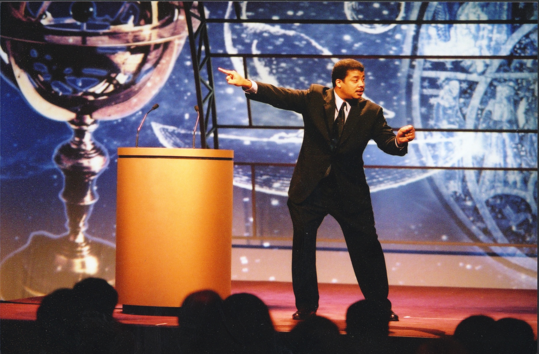 Neil deGrasse Tyson expressively pointing into the air while on stage during a speech.