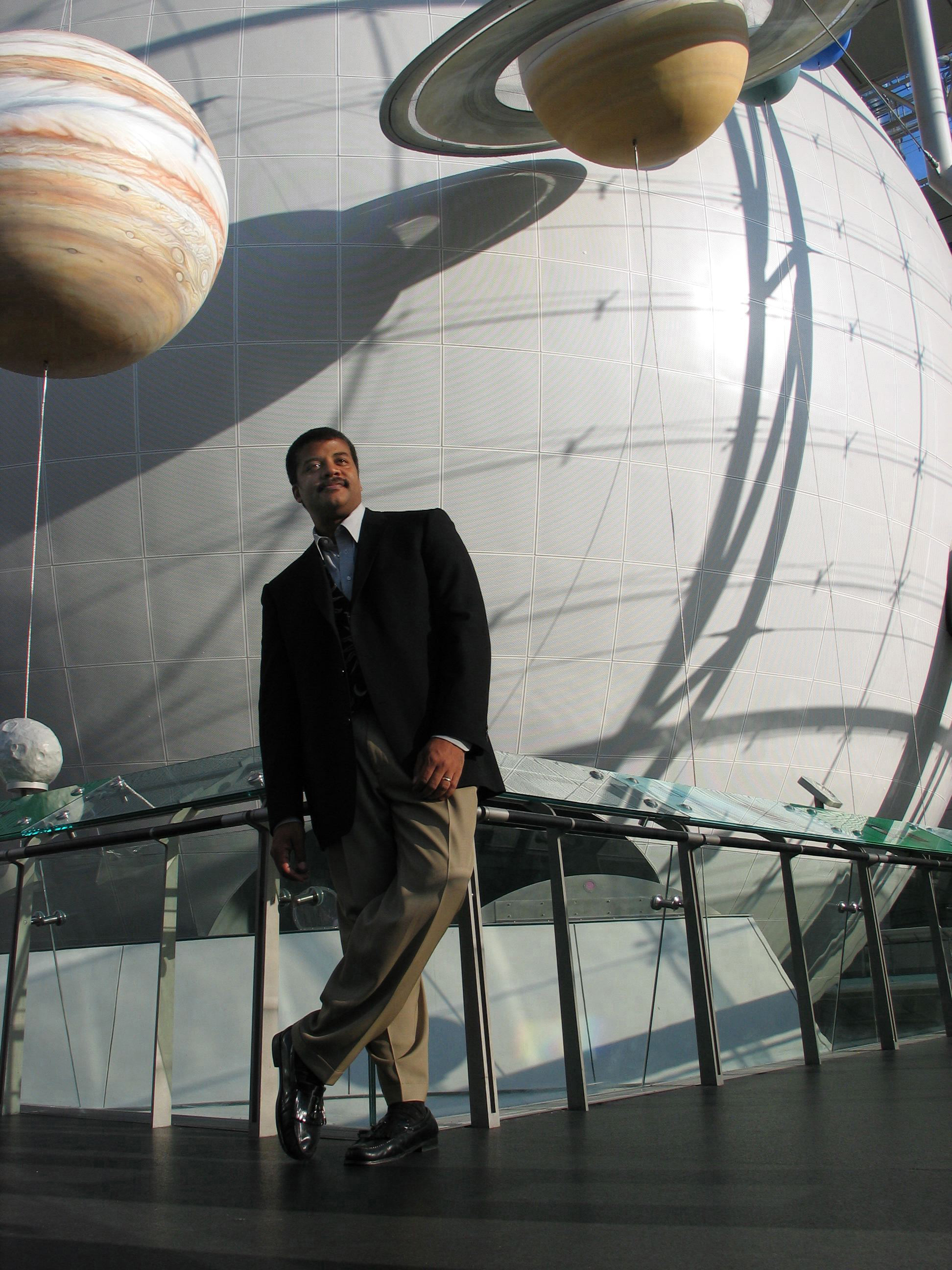 Neil deGrasse Tyson standing in the Rose Center for Earth and Space with the huge sphere behind him, and the planets Jupiter and Saturn hanging beside it.
