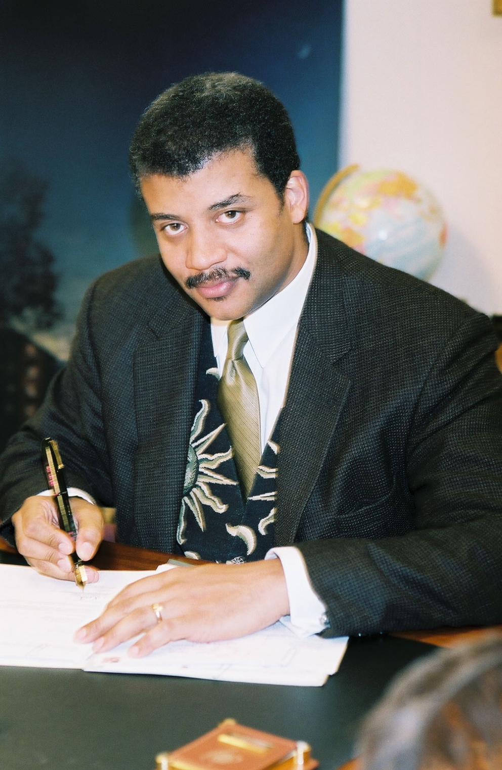 Neil deGrasse Tyson sitting at his desk signing a paper.