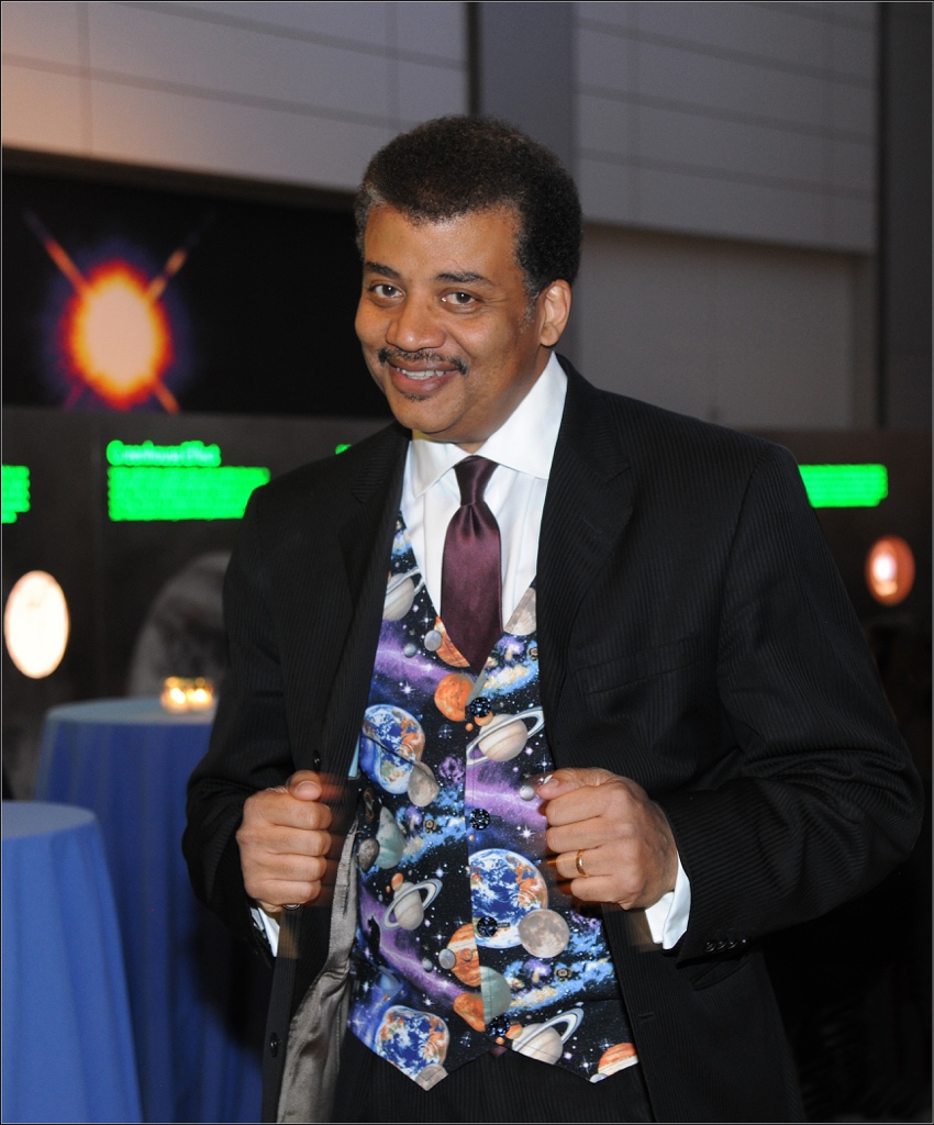 Neil deGrasse Tyson standing in the Hall of the Universe holding his jacket lapels and showing off his planet vest.