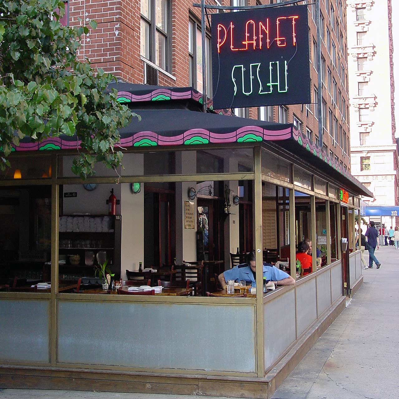 Exterior of Planet Sushi, looking down Amsterdam Avenue, with a neon sign and outdoor tables.