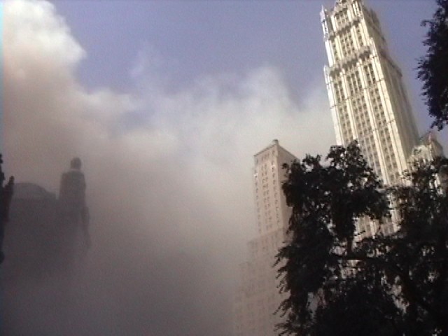 Looking toward the World Trade Center, which has collapsed, leaving a void in the sky, with thick smoke and dust in the air below.