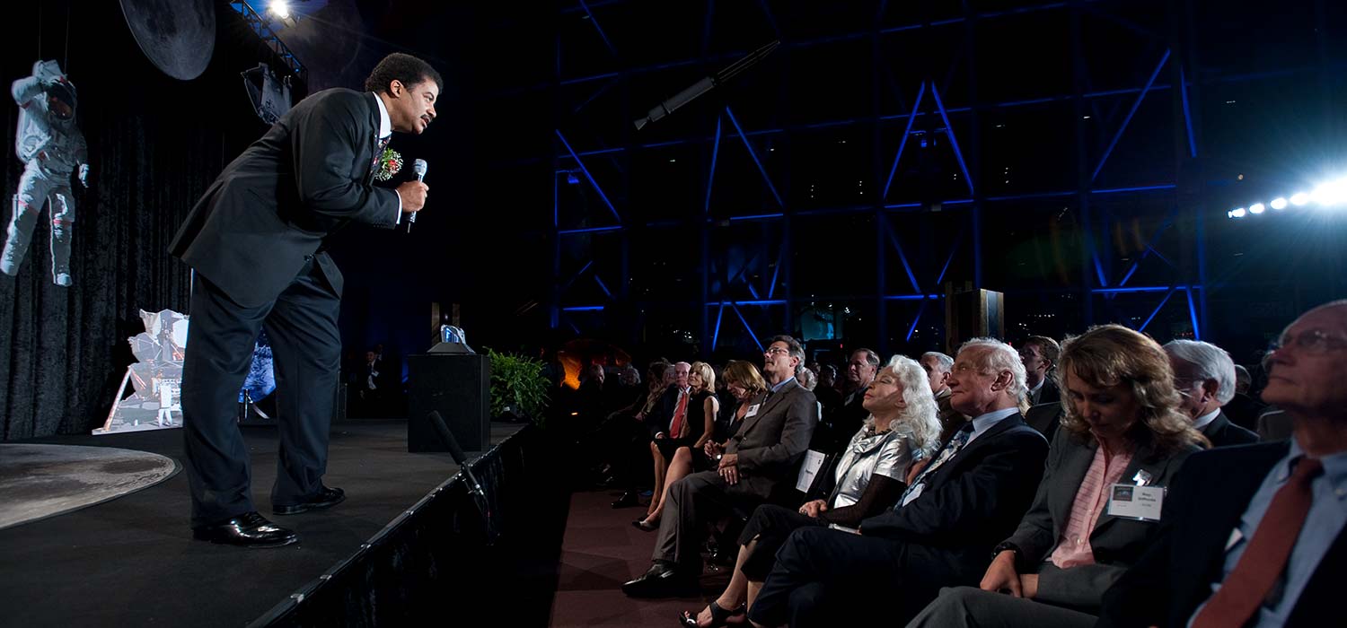 Neil deGrasse Tyson stands on the edge of a stage as he speaks to a crowd.