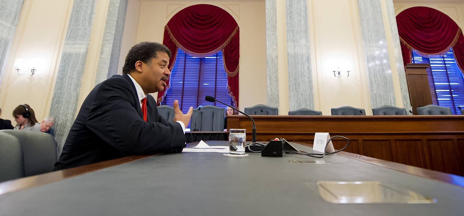 Neil deGrasse Tyson testifying before the U.S. Senate, seated at a table looking toward the right, where the committee members are out of the frame.