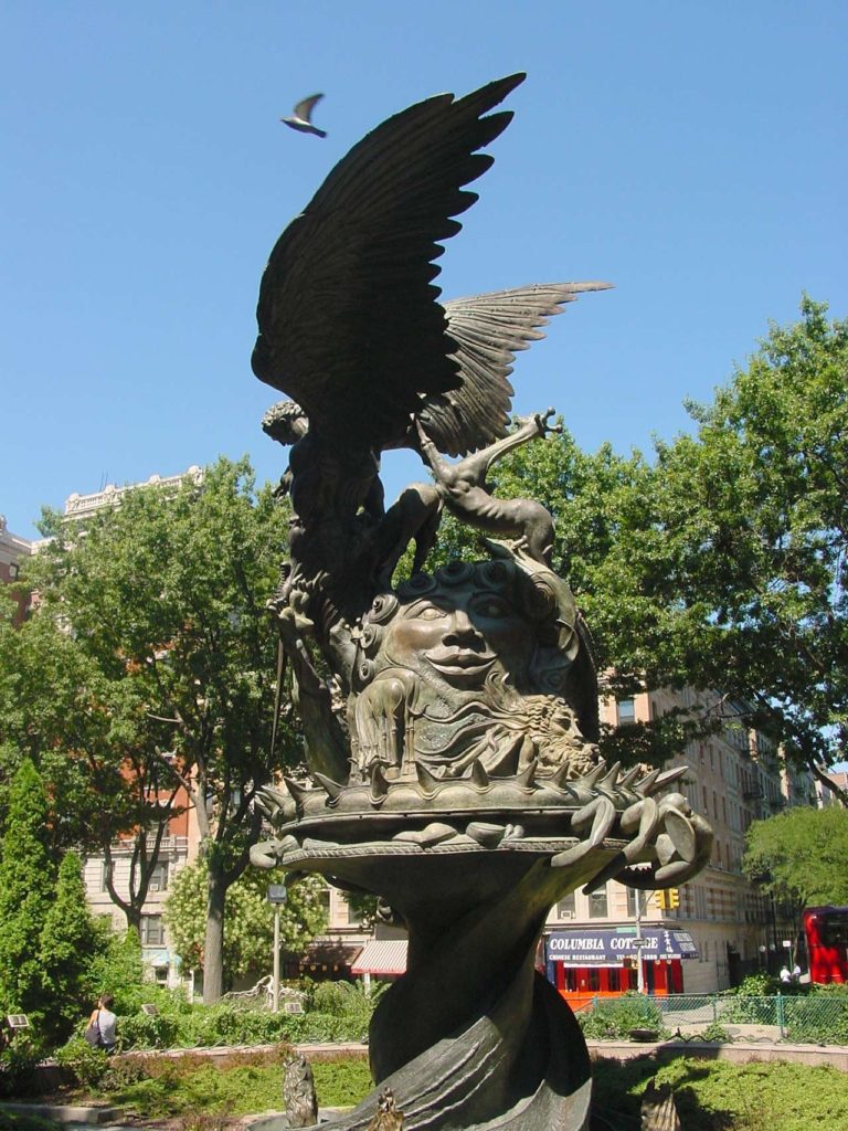 Photo of an intricate sculture resembling the centerpiece of a fountain, but there's no water running. In the center is a sun with a happy face, below a large angel with sprawling wings.