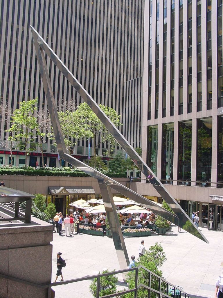 A photo of the Sun Triangle, a large, shiny, metal oblong triangle resting on one support in the plaza in front of a skyscraper.