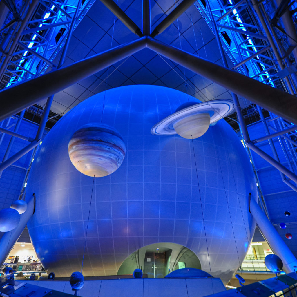 Wide angle shot inside the Rose Center, with the large sphere illuminated in blue light and Jupiter and Saturn hanging from the ceiling.